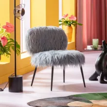 Der KARE Sessel "Hairy" in Grau mit flauschigem Kunstfellbezug und schlanken, schwarzen Metallbeinen steht in einem farbenfrohen Raum. Im Hintergrund befinden sich gelbe Wände, grosse Fenster mit rosa Vorhängen sowie gr