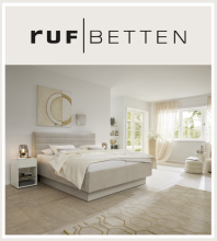 Modernes Schlafzimmer mit RUF Boxspringbett RUF Betten Logo mit beigem Boxspringbett in hellem, modernem Schlafzimmer mit Fenstern und Sitzecke.