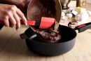 Person füllt Schokoladenteig mit rotem Silikonspatel in schwarze Backform, Backen, Küche, Zubereitung.