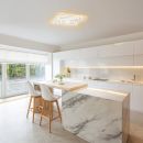 Moderne, helle Küche mit Marmorinsel, LED-Beleuchtung, weißen Hochglanzfronten und großen Fenstern.