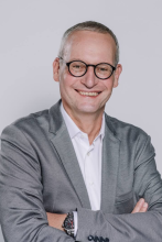 Porträt von Benno Rieger, freundlich lächelnd, mit Brille und grauem Sakko über weissem Hemd, die Arme verschränkt vor einem neutralen Hintergrund – professionelles Businessfoto.