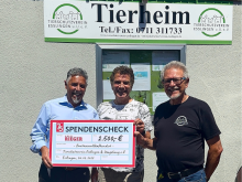 rei Männer übergeben vor einer Wand einen Spendenscheck an das Tierheim Heilbronn, mit Logo im Hintergrund.