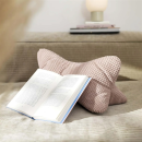 Rosa Nackenkissen aus Strickstoff auf Sofa, davor aufgeschlagenes Buch, gemütliche Leseecke im Wohnzimmer.