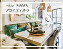 Modern eingerichteter Essbereich mit grossem Hochglanztisch, dekorativen Kissen und Hängelampen, oben links Text „Möbel Rieger Wohnstyling“.  
