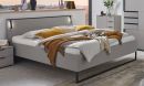 Bett Tilburg Saharagrau 180 x 200 cm Modernes Schlafzimmer mit grauem Boxspringbett, gelben Kissen, Tagesdecke und stilvoller Dekoration bei Sonnenuntergang.