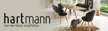 Werbebanner der Marke Hartmann mit dem Slogan „Von der Natur empfohlen“. Gezeigt wird ein moderner Essbereich mit Holztisch und schwarzen Stühlen, am Boden sitzt ein Dachs zwischen herbstlichem Laub.