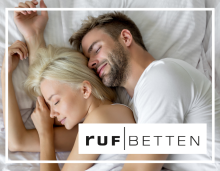 RUF Betten Werksberatung Paar liegt entspannt kuschelnd im Bett, unten rechts das Logo von ruf Betten.