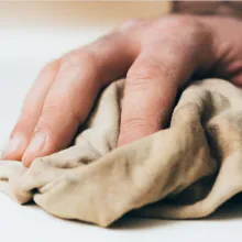 Nahaufnahme einer Hand, die mit einem beigefarbenen Tuch eine Oberfläche wischt – Symbol für Reinigung oder Pflege von Möbeln.