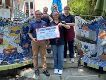 Gruppe von Menschen übergibt symbolischen Spendenscheck vor einer Wand mit buntem Mosaikdesign.